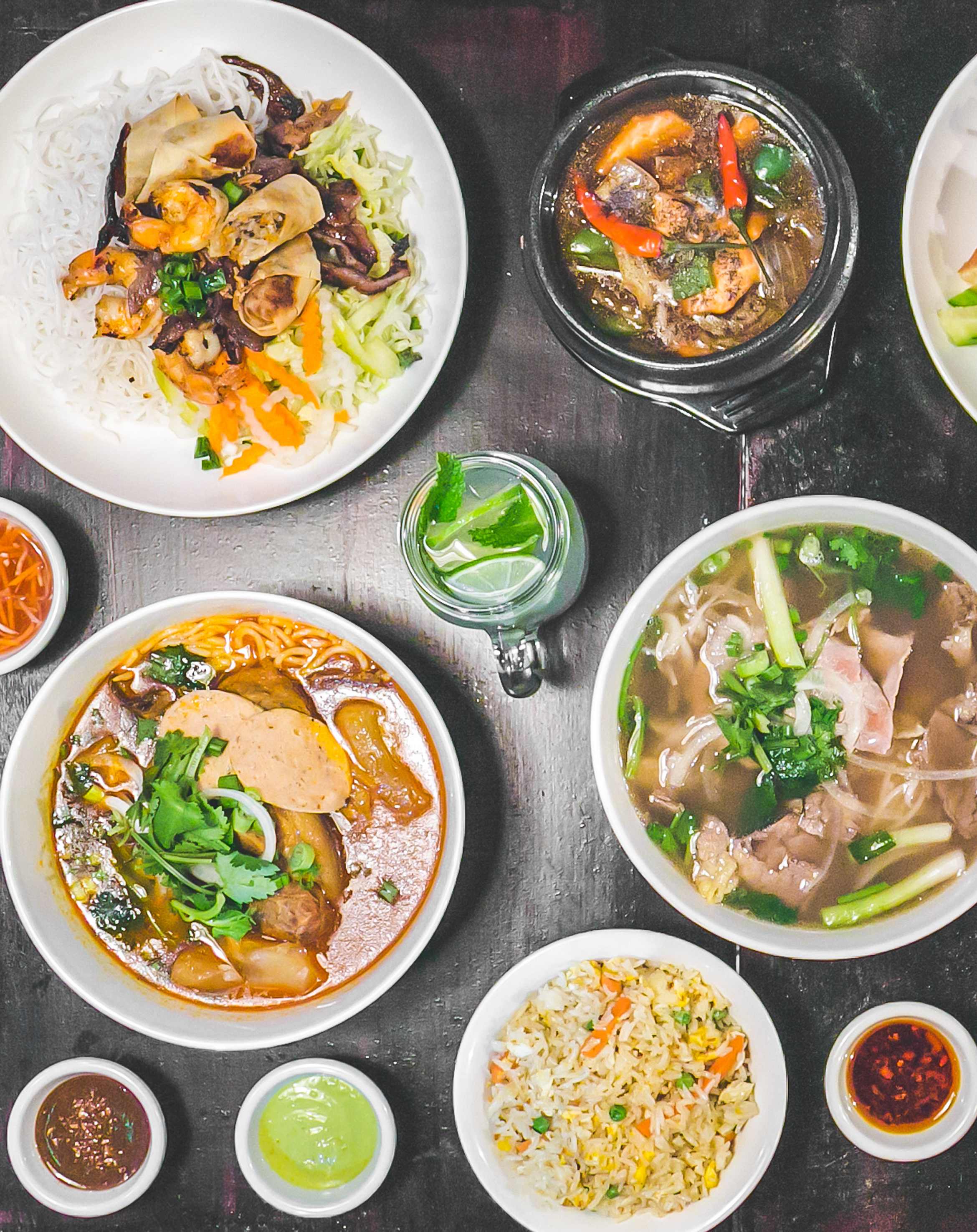 Overhead spread of phở, bún bò huế, clay pot, and spring rolls on dark wood
